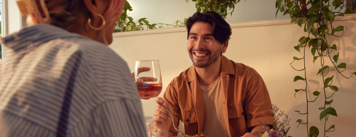 A smiling person holding a glass of drink, sitting at a table with another person, with green plants and a light-colored wall in the background.