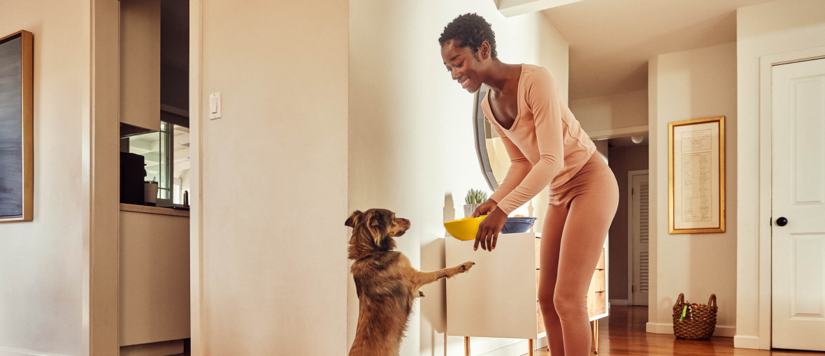 A woman in pink loungewear smiling and feeding her dog inside a modern, cozy home.